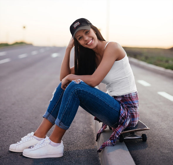 Girl wearing black dad cap with SBK9 log sitting on skateboard by curb wearing blue jeans, white tank top and shoes